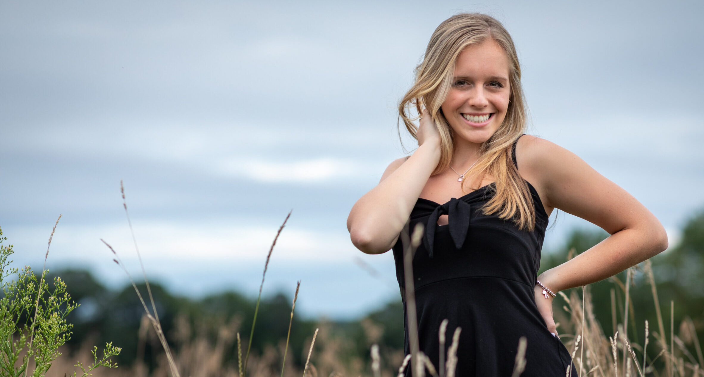CT Senior Photographer | CT Photographer | West Hartford Senior Portraits | Smiling HS Senior in a black dress standing in a field of tall grass under a cloudy sky, photographed by Rob Faber Photography.