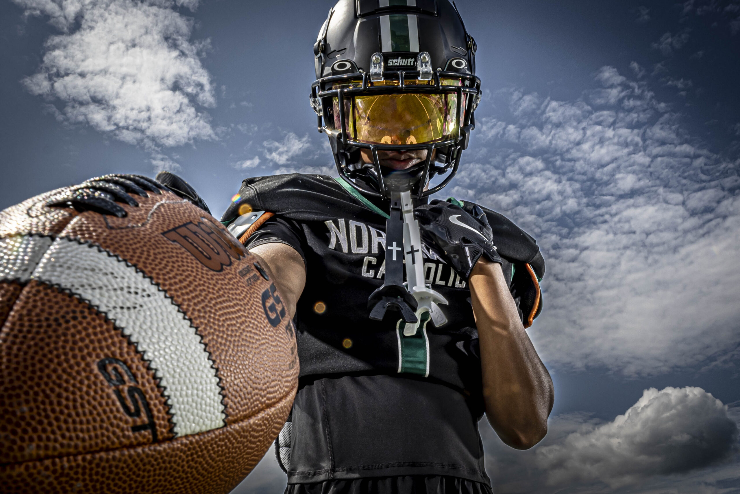 Senior sports portrait in CT of a high school football player in black uniform and helmet, holding a football toward the camera under a blue sky.