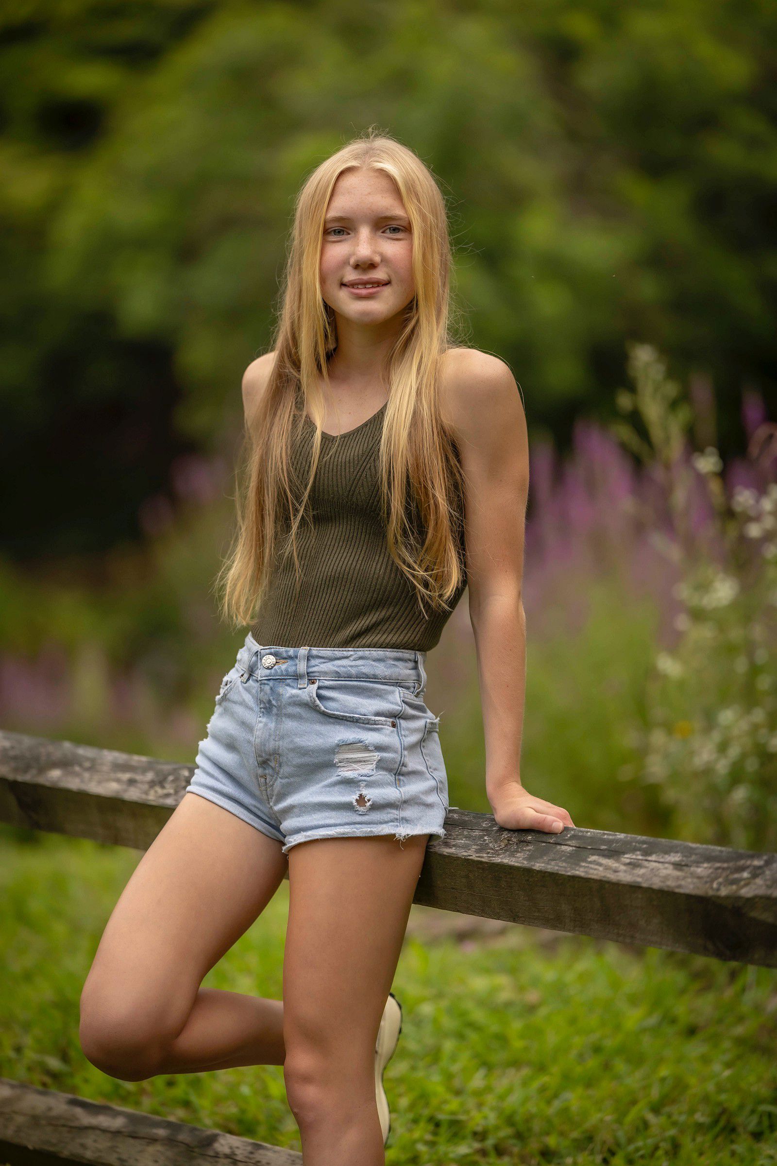 Senior portrait of a female high school senior leaning against a rustic wooden fence, wearing a neutral tank top and denim shorts, photographed outdoors with soft greenery and natural light for a relaxed, authentic look.