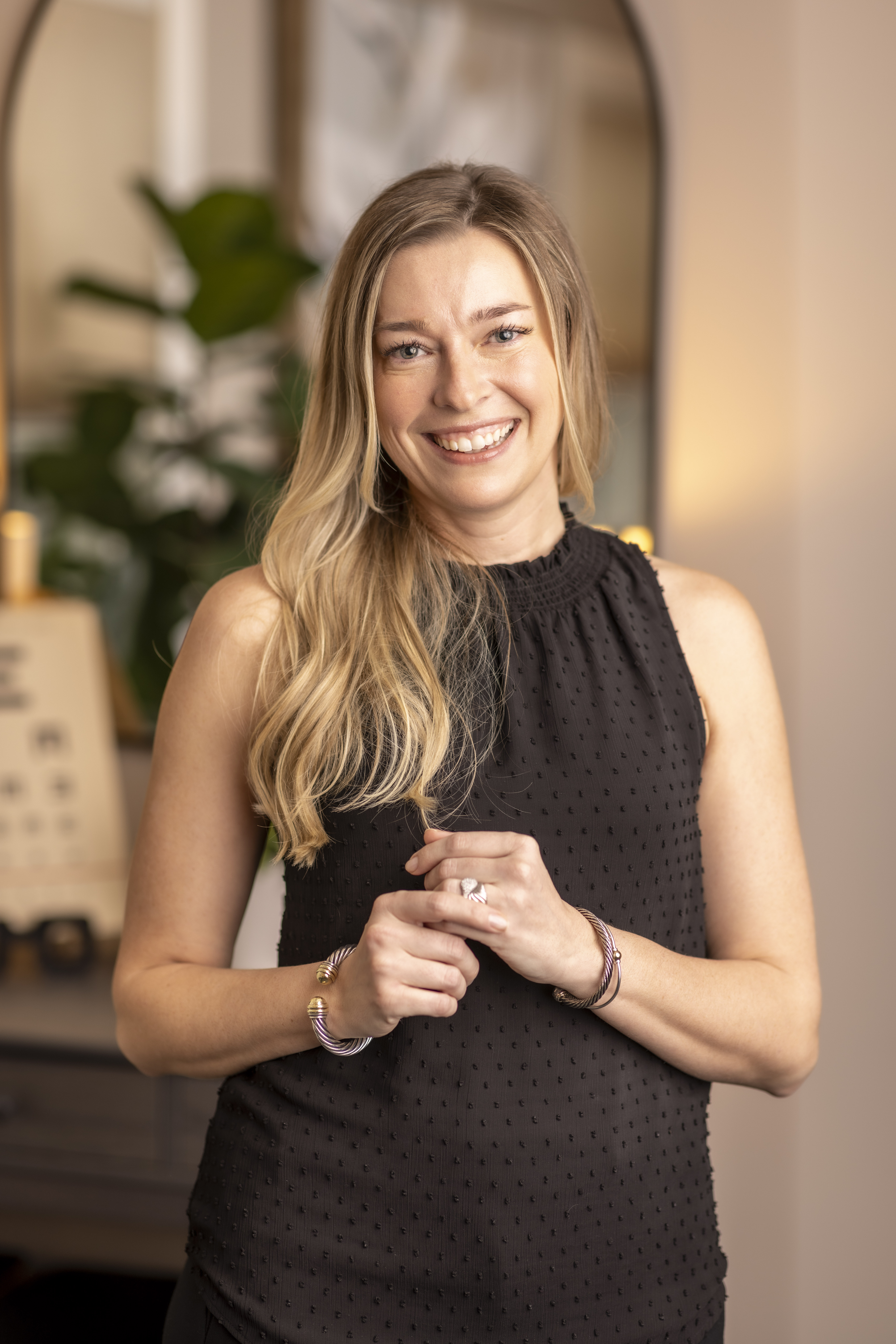 Professional headshot of a smiling optometrist wearing a sleeveless black top, standing indoors with hands gently clasped, photographed in soft natural light with a clean, modern background that conveys approachability and professionalism.
