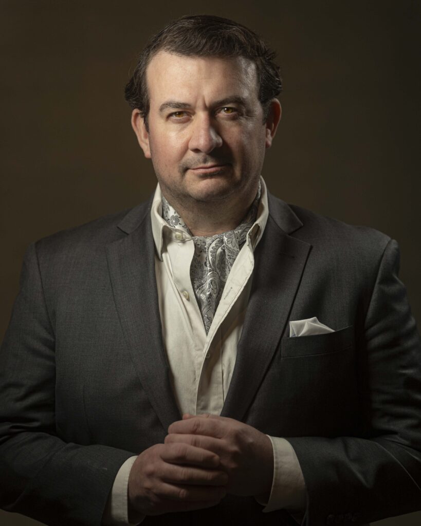 Professional headshot of a man in a dark suit jacket and white shirt, hands clasped, photographed with dramatic studio lighting against a dark background, conveying confidence and professionalism.