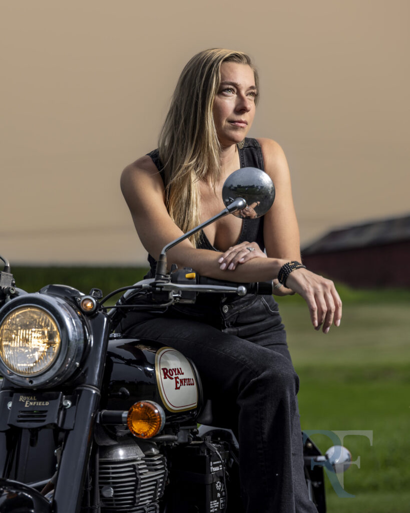A woman with long blonde hair, wearing a black jumpsuit, leans on the handlebars of a vintage Royal Enfield motorcycle. She gazes into the distance against a softly lit sky, showcasing a cinematic fine-art portrait session by a CT Photographer in Northern Connecticut.