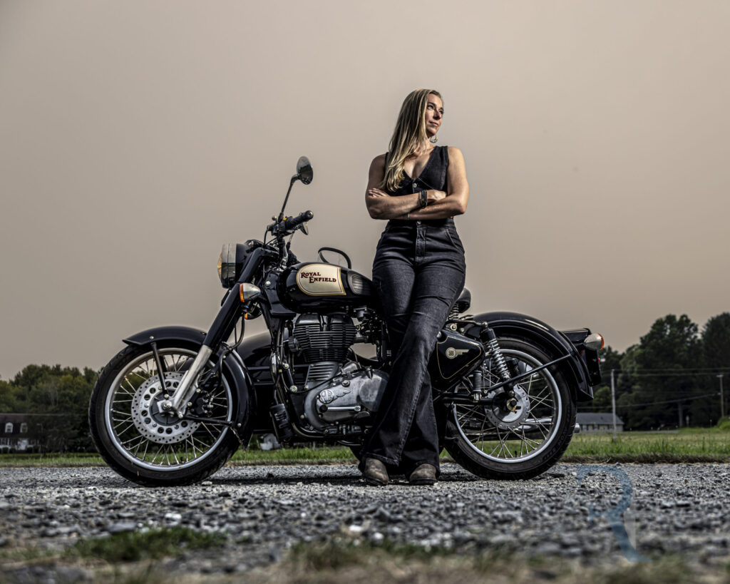 Avon Photographer | Hybrid Session| Woman donning a black jumpsuit, stands with arms crossed beside a vintage black Royal Enfield motorcycle on a gravel road at sunset in rural Connecticut. The dramatic lighting and moody sky create a bold, fine-art portrait captured by CT Photographer Rob Faber.
