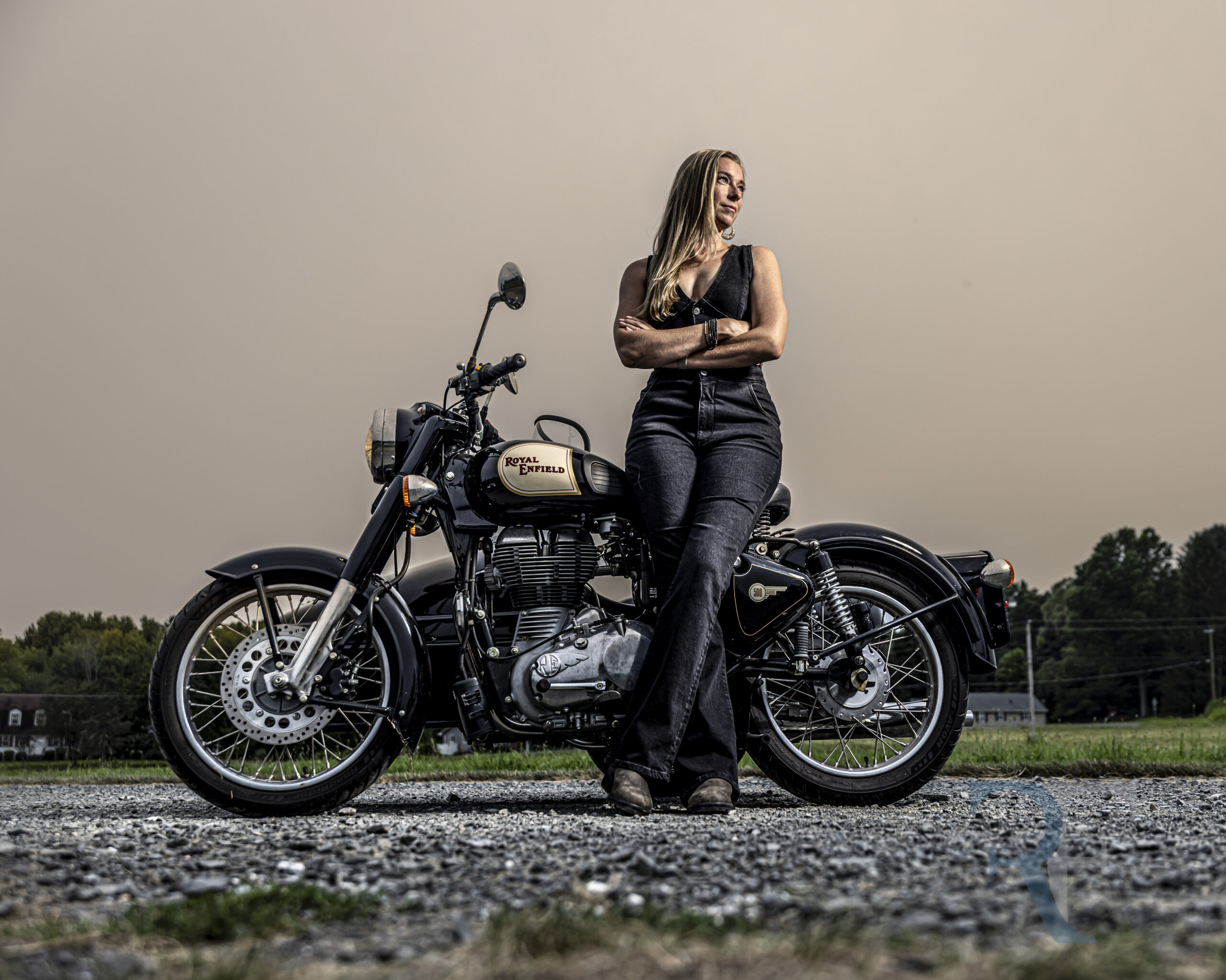 Avon Photographer | Hybrid Session| Woman donning a black jumpsuit, stands with arms crossed beside a vintage black Royal Enfield motorcycle on a gravel road at sunset in rural Connecticut. The dramatic lighting and moody sky create a bold, fine-art portrait captured by CT Photographer Rob Faber.