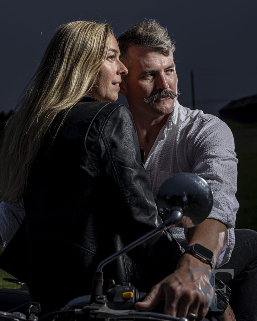 Backlit close-up of a couple on a Royal Enfield motorcycle, the woman in a black leather jacket and the man steering, captured in cinematic lighting. This romantic and edgy image exemplifies luxury portrait photography in CT and Western Mass.