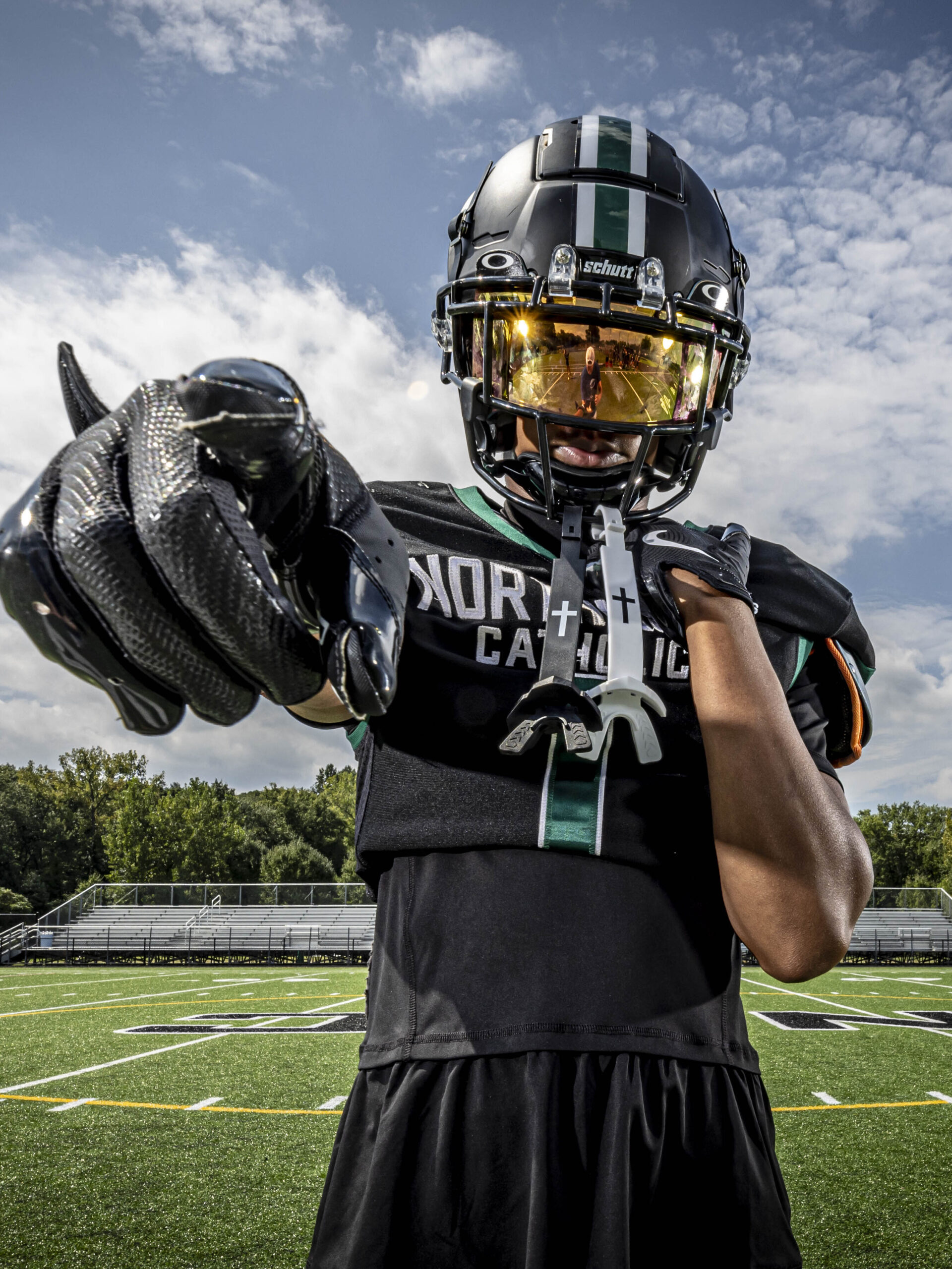 Senior sports portrait in CT of a high school football player in black uniform and helmet, holding a football toward the camera under a blue sky.