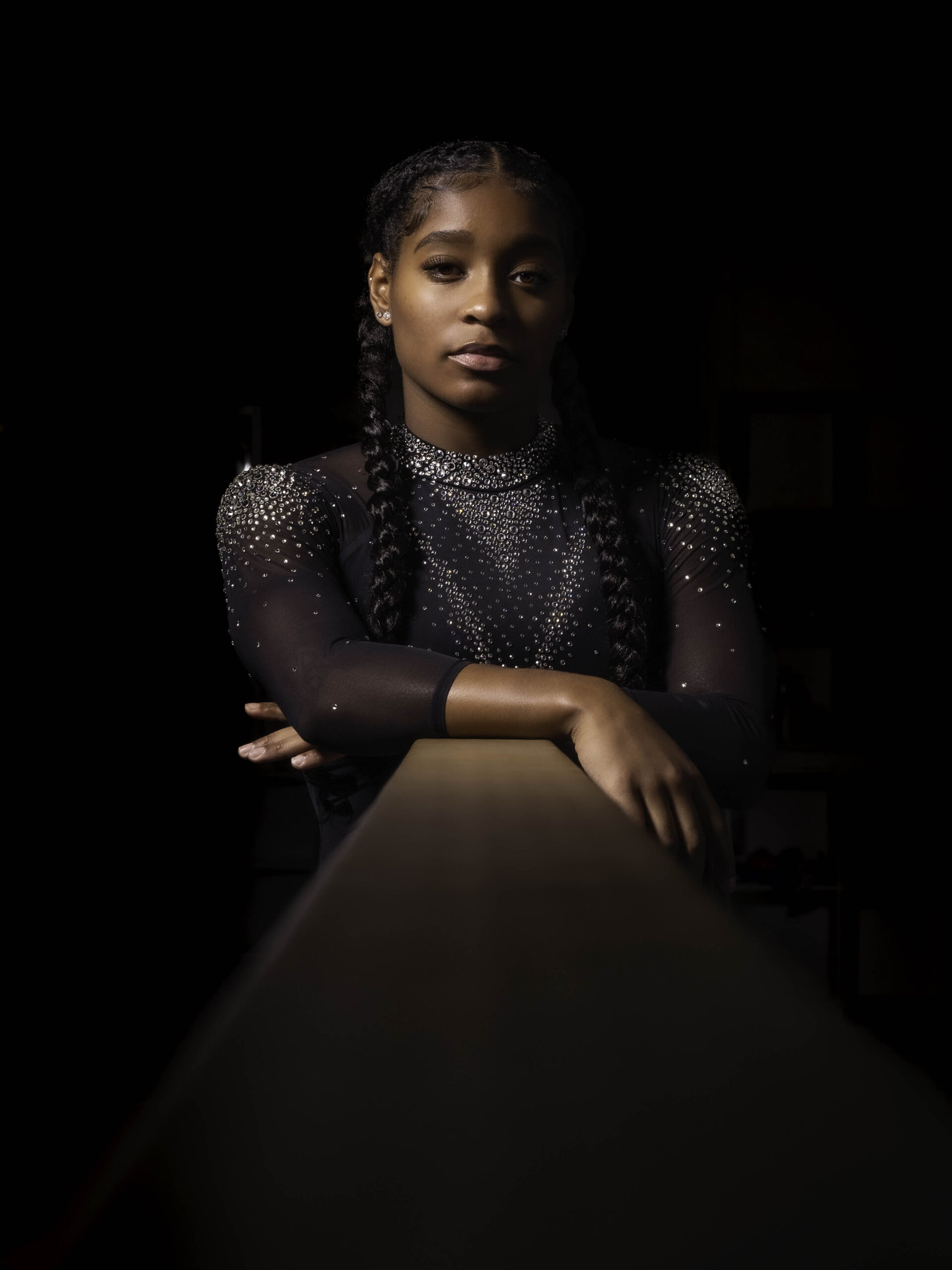 Senior portrait in CT of a young woman with long braids, wearing a sparkly black top, leaning on a wooden surface against a dark background.
