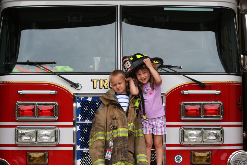 Photographer in CT | CT Photographer | Suffield Photographer captures two children smiling in front of a fire truck, dressed in oversized firefighter gear and helmet, showcasing a family moment connected to firefighting service.