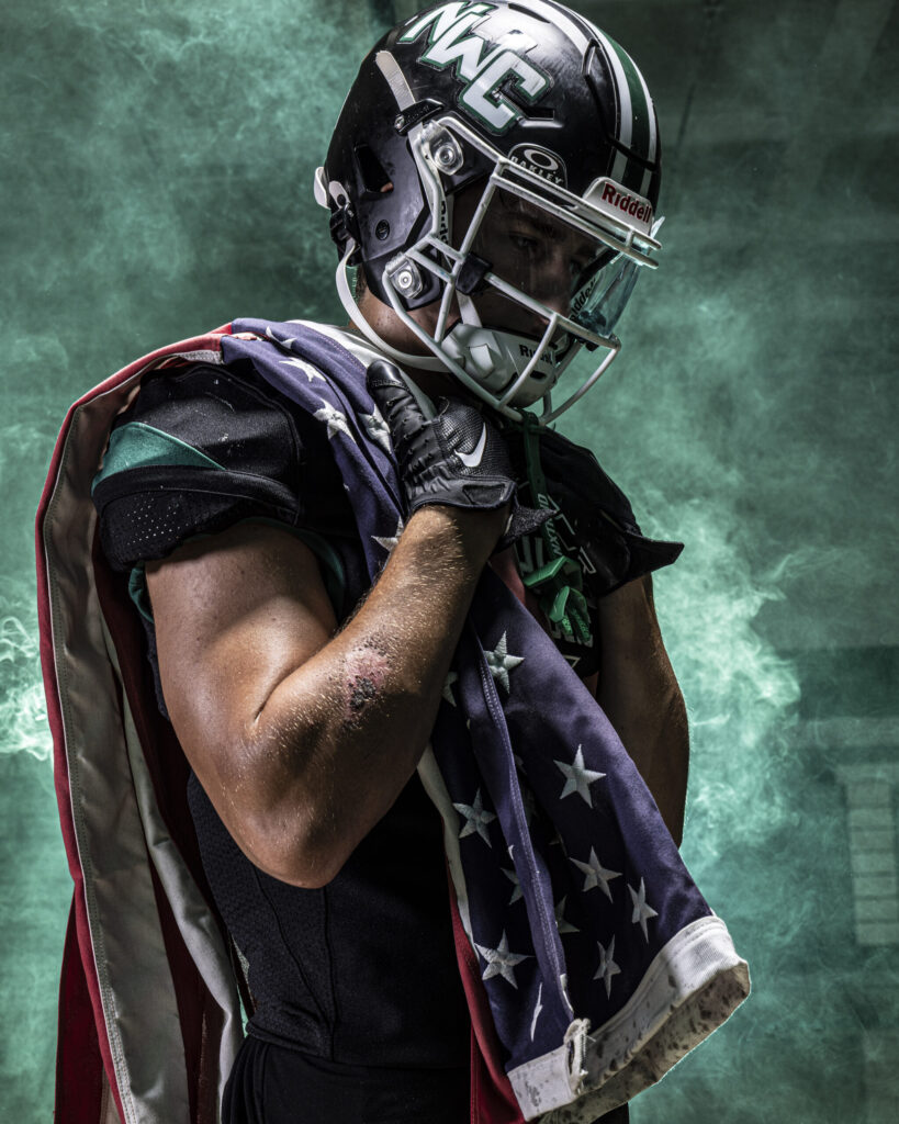CT Sports Photographer | CT Photographer | Rob Faber Photography - Football player draped in an American flag looks down with focus, helmet on, green smoke swirling in the background.