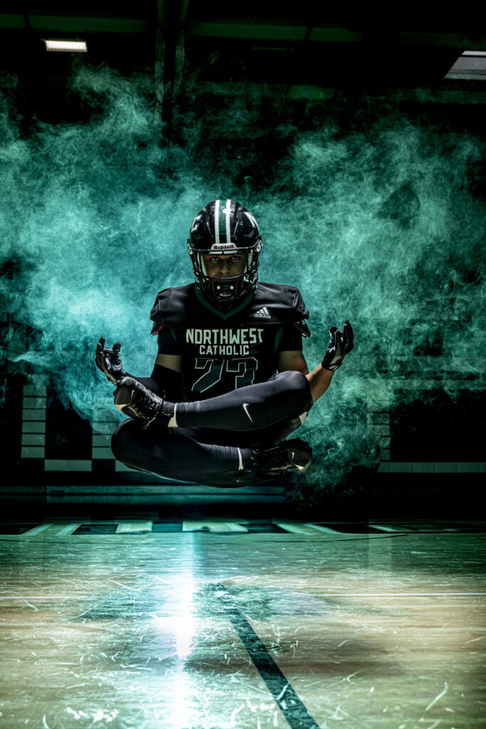 CT Sports Photographer | CT Photographer | Rob Faber Photography - Football player levitates in lotus pose with helmet on, centered on court with teal smoke behind, creating a surreal and cinematic sports portrait.