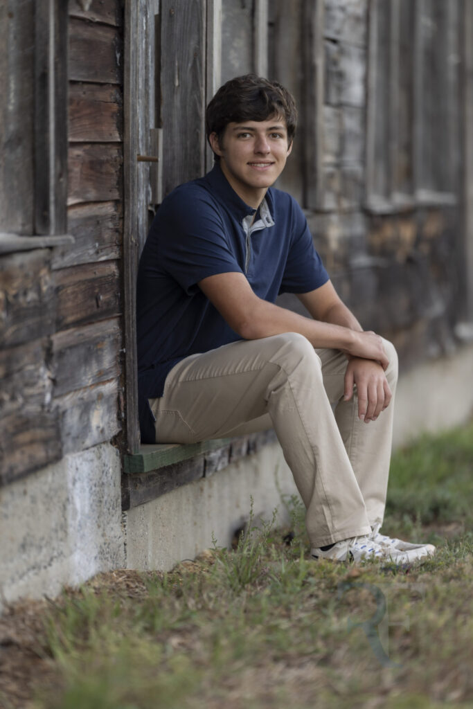 Photographer in Simsbury | Rob Faber Photography | Senior portrait of Thomas sitting by a rustic wooden barn wall in a navy polo and khaki pants, captured in natural light for a timeless outdoor session.