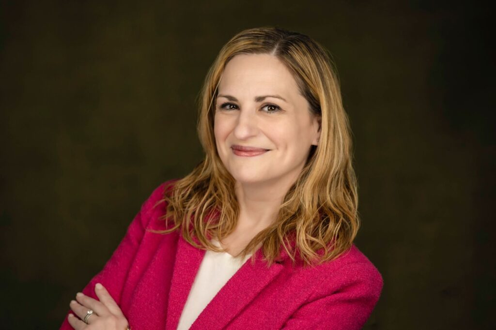 Warm professional portrait of a woman in a pink blazer and white top, photographed against a dark green studio backdrop in Connecticut.