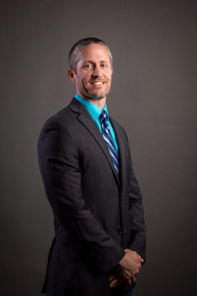 Professional portrait of a smiling man wearing a charcoal suit and teal shirt with blue tie against a gray background, photographed in CT studio.
