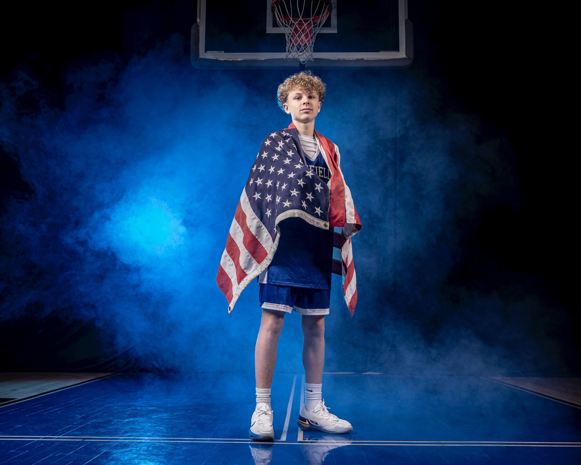 Rob Faber Photography | Media Day Photographer | What is a Media Day | High school basketball player standing on a court wrapped in an American flag, photographed during a professional media day with dramatic blue lighting and smoke to convey confidence, pride, and athletic presence.