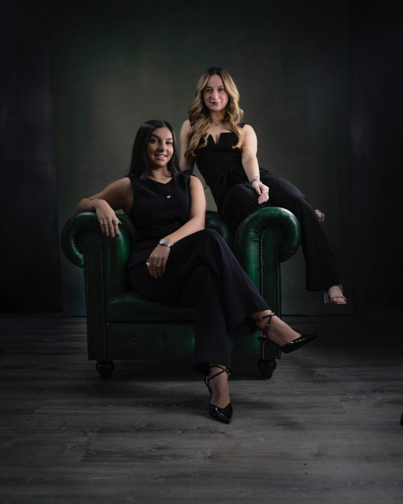 Rob Faber Photography | Headshot Photos | Professional branding portrait of two young women seated on a green leather chair in a studio, wearing black outfits and posing confidently under dramatic studio lighting.