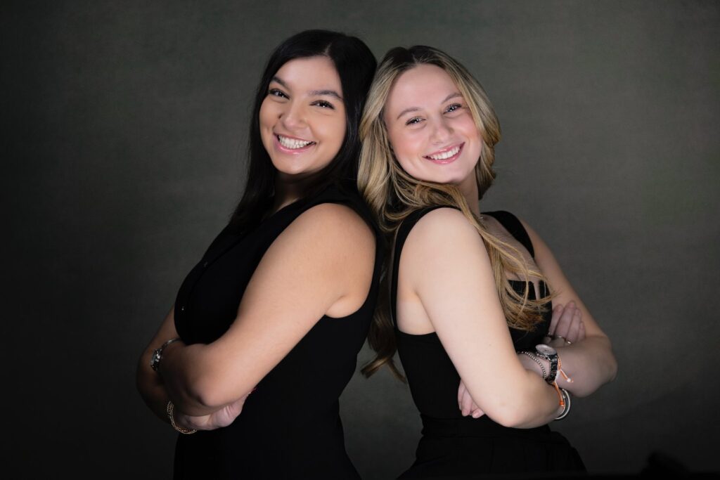 Rob Faber Photography | Headshot Photos | Professional headshots in CT of two young women standing back to back in a studio, smiling confidently in coordinated black outfits against a dark background.