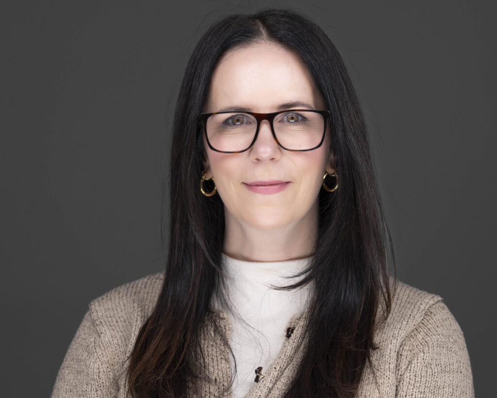 Rob Faber Photography | CT Photographer | Professional studio headshot of a woman with long dark hair and glasses wearing a cream sweater and gold hoop earrings against a dark gray background, photographed with soft modern lighting for business branding and professional profile photography.
