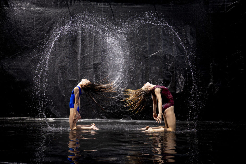 Rob Faber Photography | CT Photographer | Rain Photos | Dance Photos | Two dancers in blue and maroon leotards kneel facing each other, flinging their long wet hair sideways so the strands and water spray meet in the center against a dark backdrop.