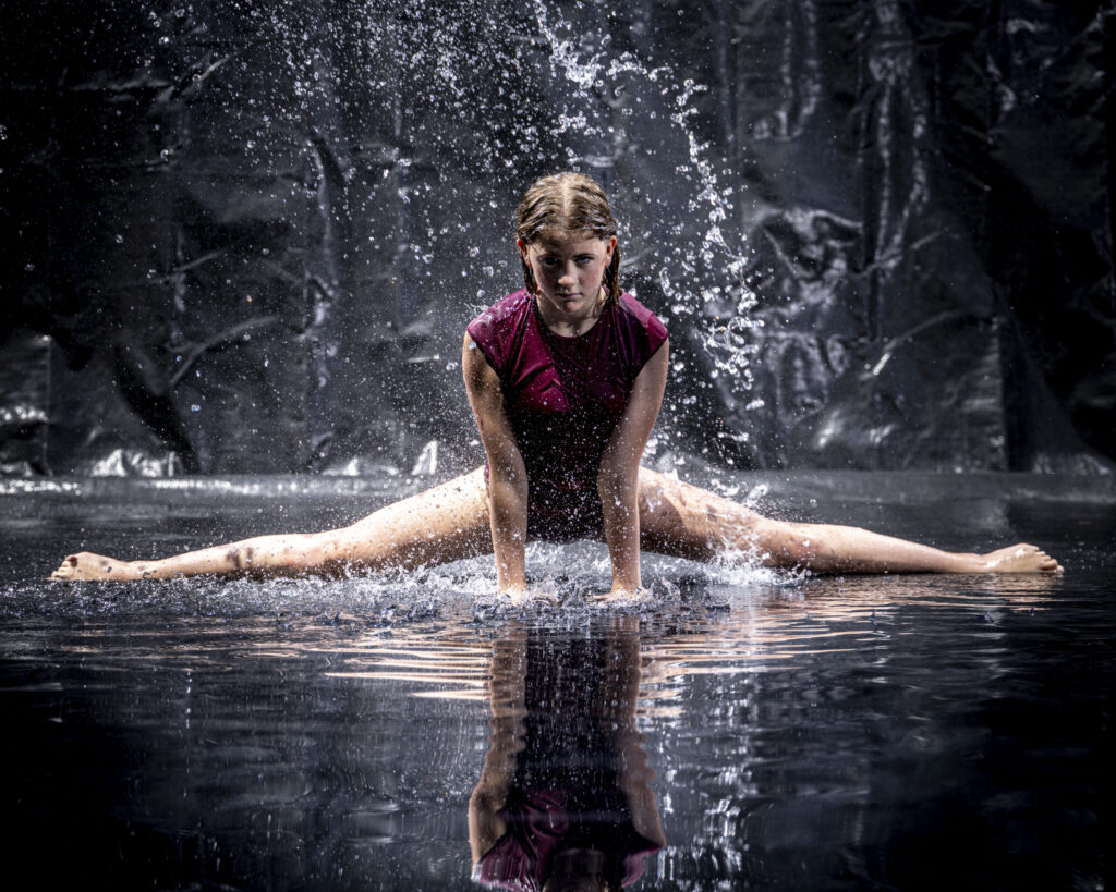 Rob Faber Photography | CT Photographer | Rain Photos | Dance Photos | Dancer in maroon holds a center split low over a wet reflective floor, gazing at the camera as water sprays up around her against a dark background.
