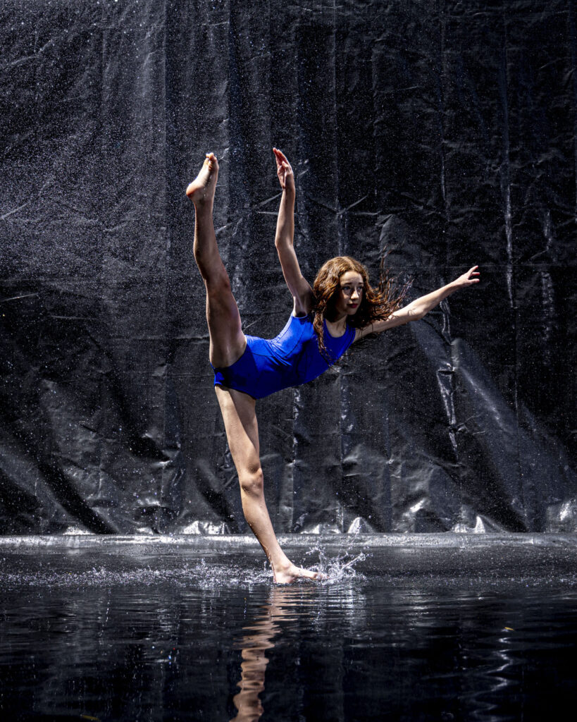 Rob Faber Photography | CT Photographer | Rain Photos | Dance Photos | Dancer in a blue leotard balances on one leg in a standing split, one arm extended, foot splashing the wet floor as water rains down against a dark background.