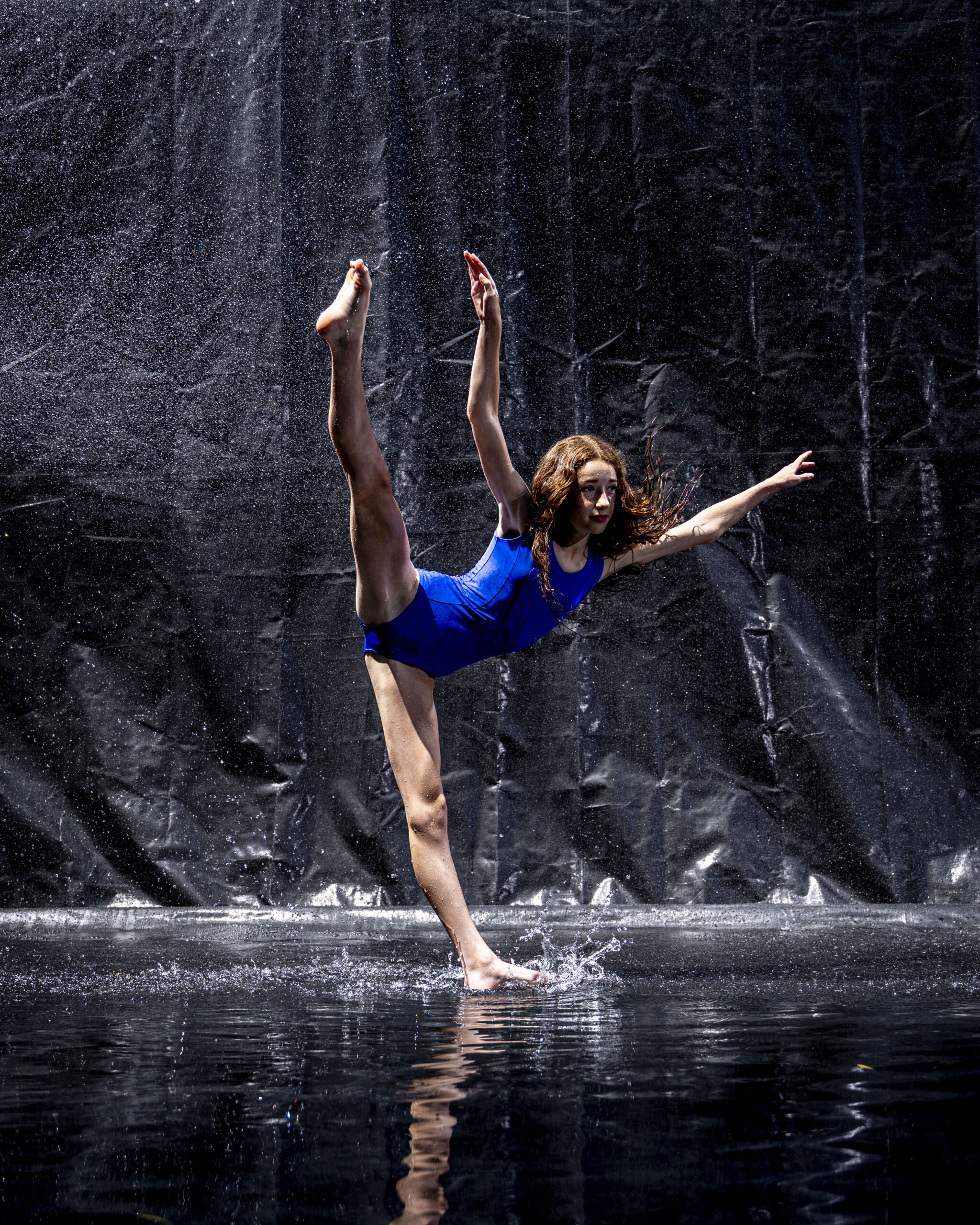 Rob Faber Photography | CT Photographer | Rain Photos | Dance Photos | Dancer in a blue leotard balances on one leg in a standing split, one arm extended, foot splashing the wet floor as water rains down against a dark background.