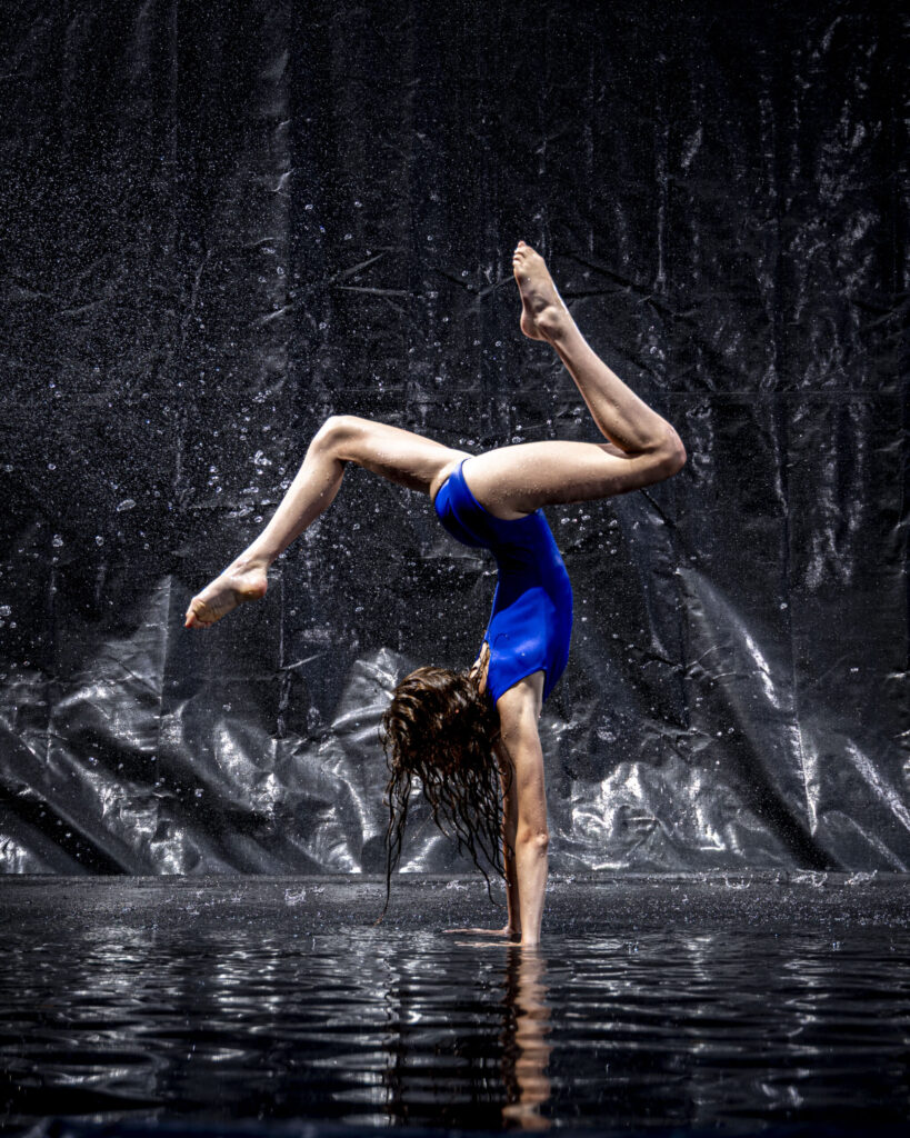 Rob Faber Photography | CT Photographer | Rain Photos | Dance Photos | Dancer in a blue leotard holds a one-handed handstand with legs bent, wet hair hanging toward the reflective floor as water sprays against a black backdrop.