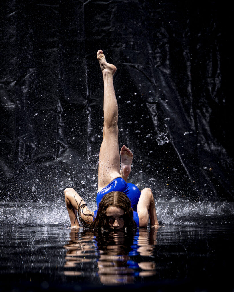 Rob Faber Photography | CT Photographer | Rain Photos | Dance Photos | Dancer in a blue leotard lies on the wet floor with legs extended straight up, gazing at the camera as water splashes around her against a dark background.
