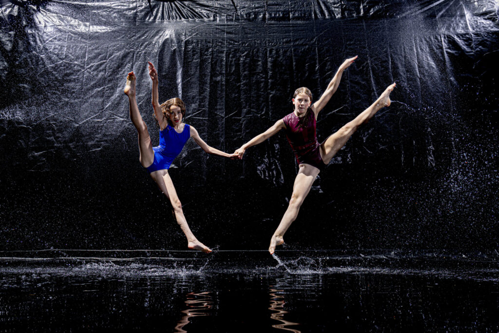 Rob Faber Photography | CT Photographer | Rain Photos | Dance Photos | Two dancers in blue and maroon leotards leap side by side in matching splits, holding hands at center as water sprays around them against a black backdrop.