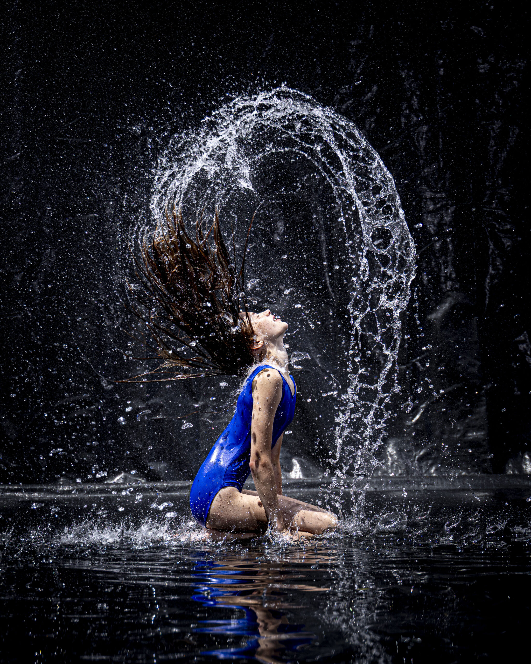 Rob Faber Photography | CT Photographer | Rain Photos | Dance Photos | Dancer in a blue leotard kneels and arches back, whipping her wet hair to send a sweeping arc of water droplets through the air against a black background.