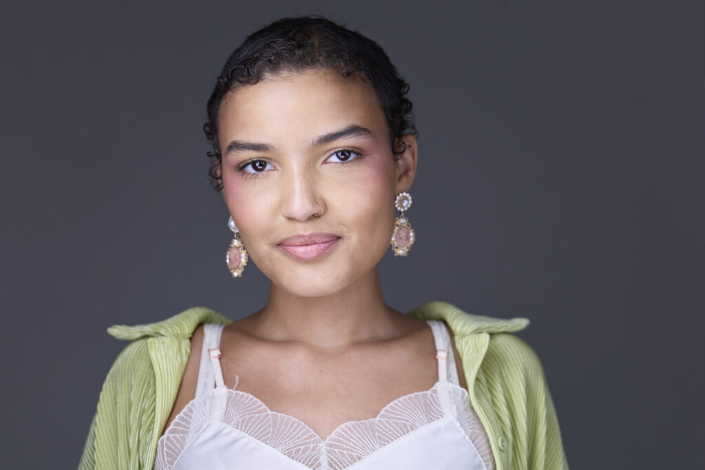CT Headshots | Rob Faber Photography | Peter Hurley Headshot Intensive | Young woman with dark curly hair pulled back, gold cameo drop earrings and a small gold nose ring, wearing a pale green pleated jacket over a white lace camisole, soft smile against a medium gray background.