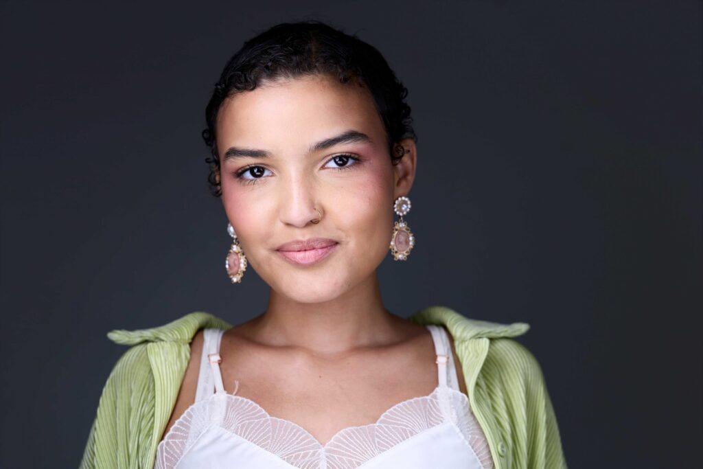CT Headshots | Rob Faber Photography | Peter Hurley Headshot Intensive | Young woman with dark curly hair pulled back, gold cameo drop earrings and a gold nose ring, wearing a pale green top over a white lace camisole, soft confident expression against a dark charcoal background.