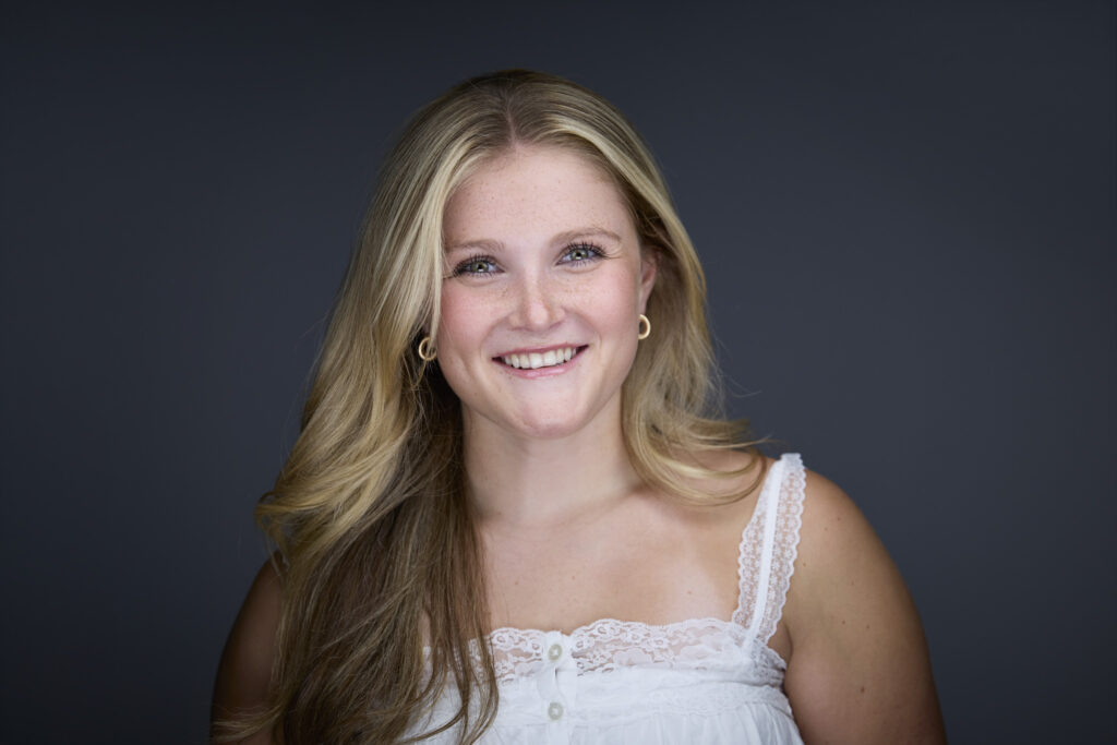 CT Headshots | Rob Faber Photography | Peter Hurley Headshot Intensive | Young woman with long blonde wavy hair and freckles, gold hoop earrings, wearing a white lace-trimmed camisole, bright cheerful smile against a dark charcoal background.