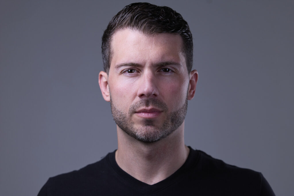 CT Headshots | Rob Faber Photography | Peter Hurley Headshot Intensive | Man with short dark hair and trimmed stubble wearing a plain black V-neck, neutral steady expression against a gray background.