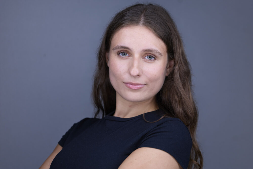 CT Headshots | Rob Faber Photography | Peter Hurley Headshot Intensive | Woman with long wavy brown hair and light eyes wearing a black short-sleeve tee, head slightly turned with a poised subtle smile against a blue-gray background.