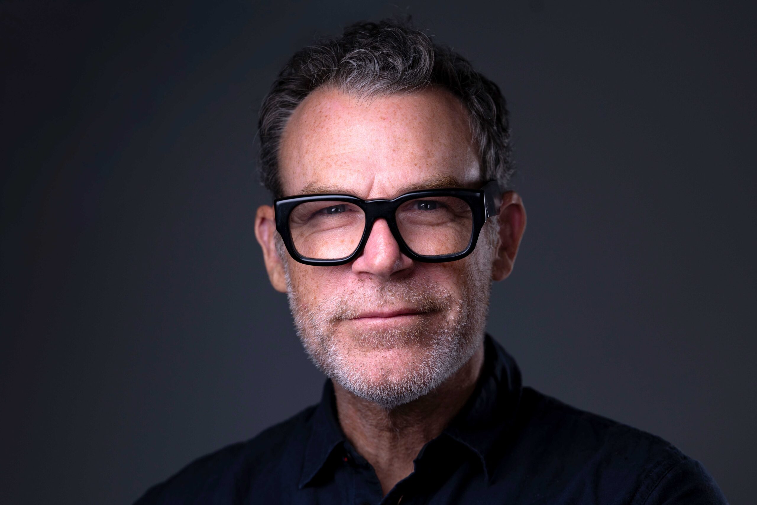 CT Headshots | Rob Faber Photography | Peter Hurley Headshot Intensive | Photographer Peter Hurley, a man with salt-and-pepper hair and a short gray beard, wearing bold black rectangular glasses and a dark button-up shirt, subtle smile against a dark gray background.