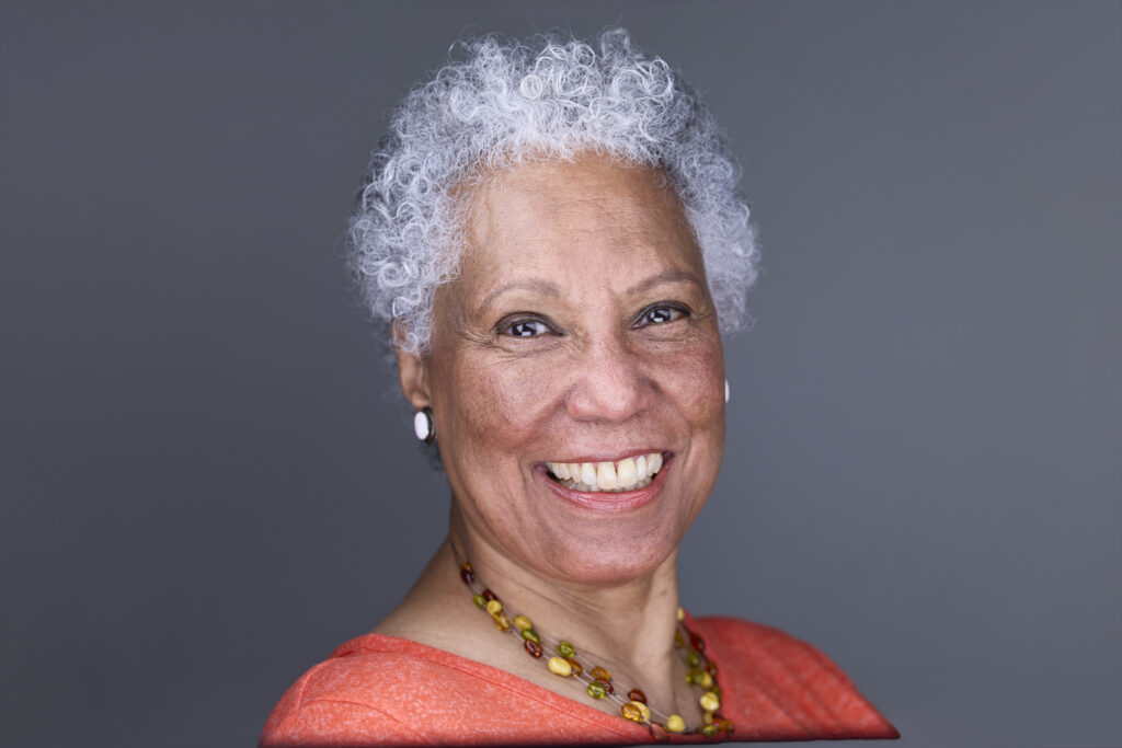 CT Headshots | Rob Faber Photography | Peter Hurley Headshot Intensive | Smiling older woman with short silver-gray curls wearing a coral top and a beaded amber necklace, warm broad smile against a gray background.