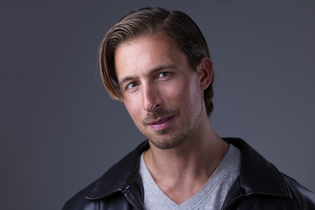 CT Headshots | Rob Faber Photography | Peter Hurley Headshot Intensive | Man with side-swept brown hair and a thin mustache and soul patch, wearing a black leather jacket over a gray V-neck tee, slight smile against a gray background.