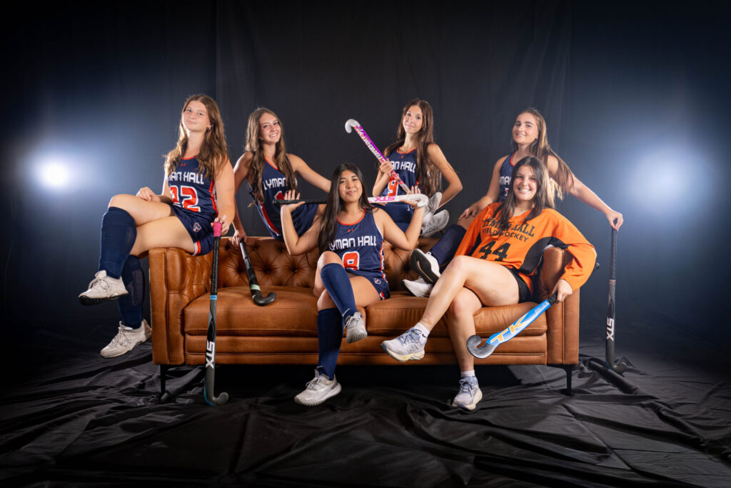 Rob Faber Photography | CT Photographer | Six Lyman Hall field hockey players posed on a brown leather tufted couch in front of a black backdrop with dramatic rim lighting, holding field hockey sticks and wearing navy and orange uniforms, with the goalie in an orange jersey.