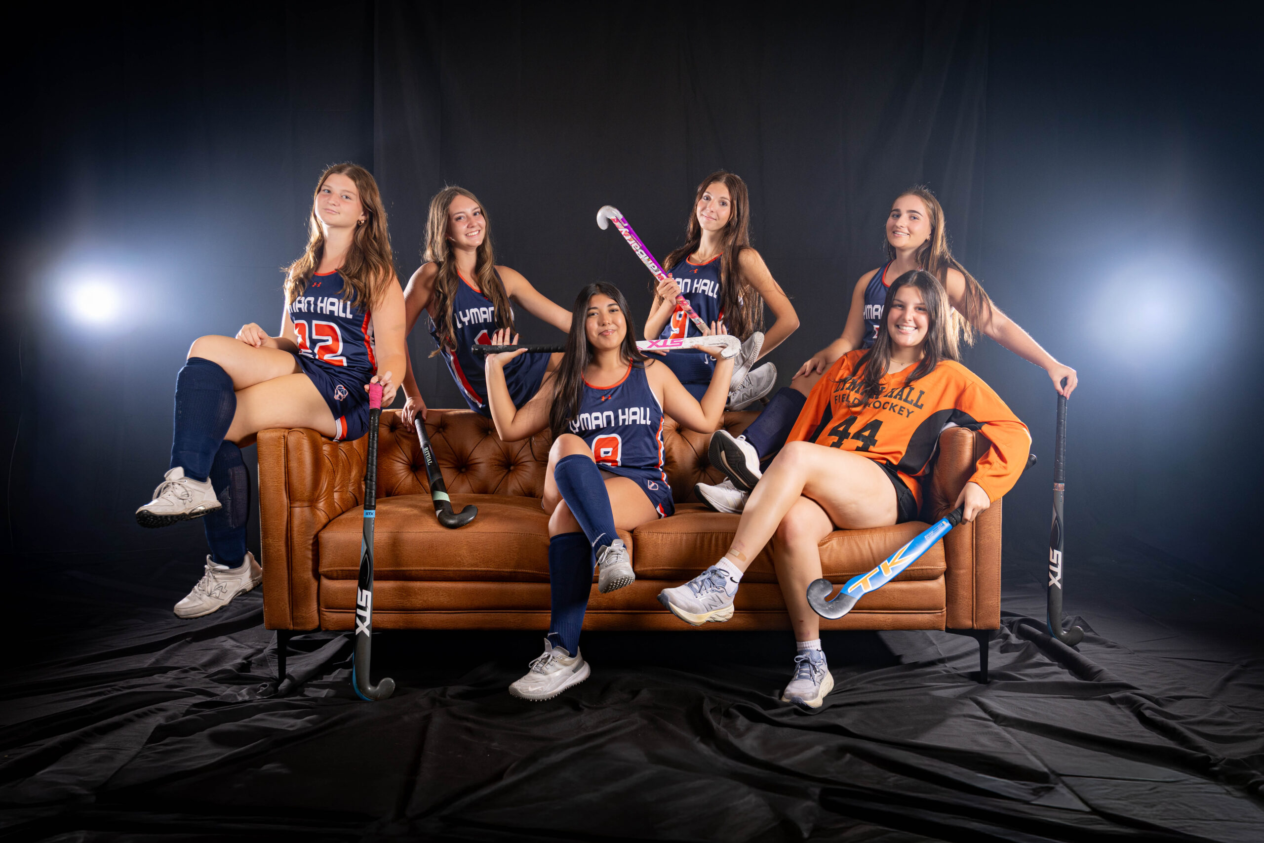 Rob Faber Photography | CT Photographer | Six Lyman Hall field hockey players posed on a brown leather tufted couch in front of a black backdrop with dramatic rim lighting, holding field hockey sticks and wearing navy and orange uniforms, with the goalie in an orange jersey.