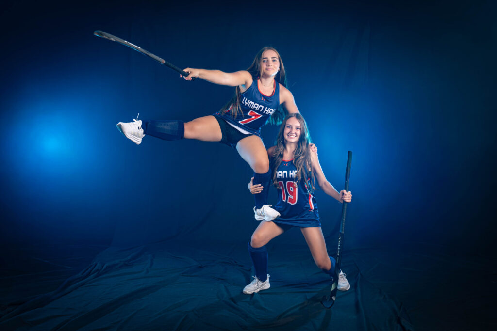 Rob Faber Photography | CT Photographer | A Lyman Hall field hockey player in a number 7 jersey performs a high kick while holding her stick overhead, supported by a smiling teammate in a number 19 jersey, against a blue-lit studio backdrop.