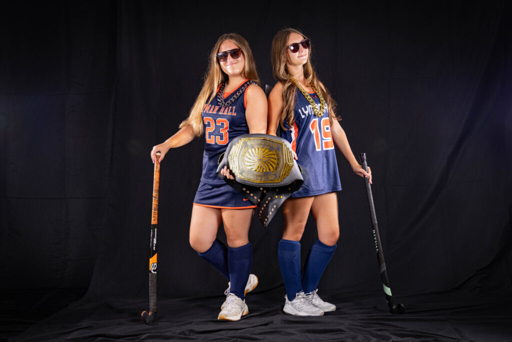 Rob Faber Photography | CT Photographer | Two Lyman Hall field hockey players in numbers 23 and 19 stand back to back wearing sunglasses and oversized chain necklaces, one holding a gold championship belt, each leaning on a field hockey stick against a black backdrop.